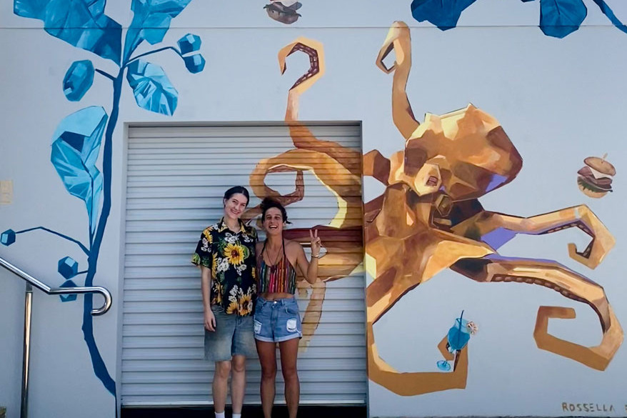 Mural artist Rossella R.Z. (right) pictured with Izzy Gabelle, daughter of the owner of Strait on the Beach, in front of the newly completed artwork that now brightens the restaurant’s exterior in Holloways Beach. Picture: Supplied